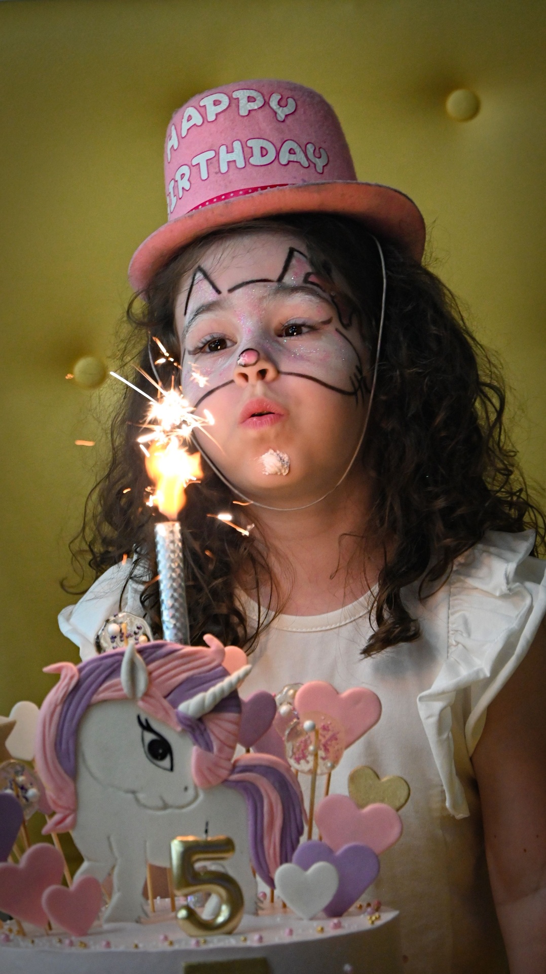 Young girl in a Happy Birthday hat blowing out a sparkler candle on her unicorn cake